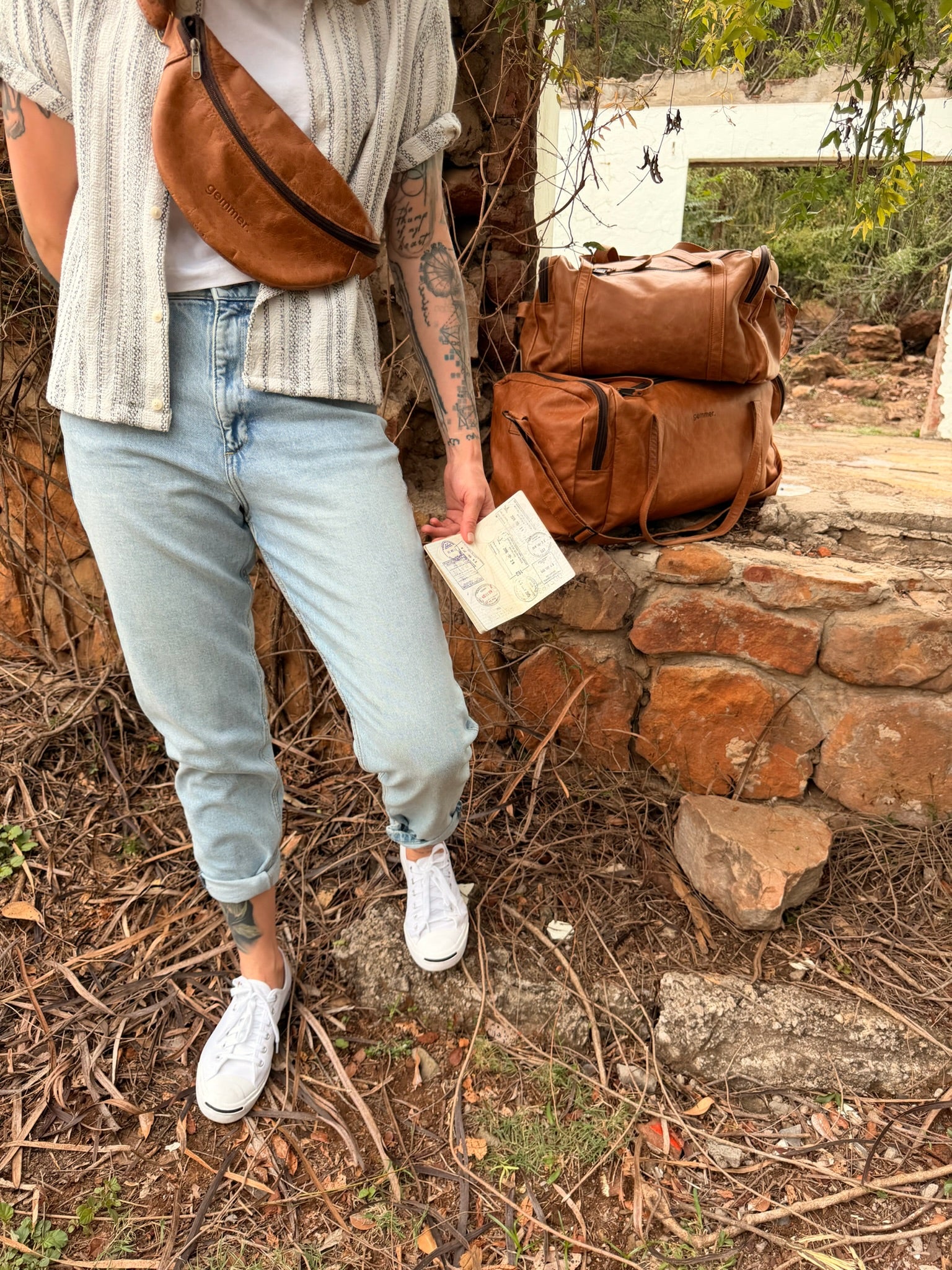 Person holding a gemmer leather bag and passport with more bags stacked in the background on a stone wall.